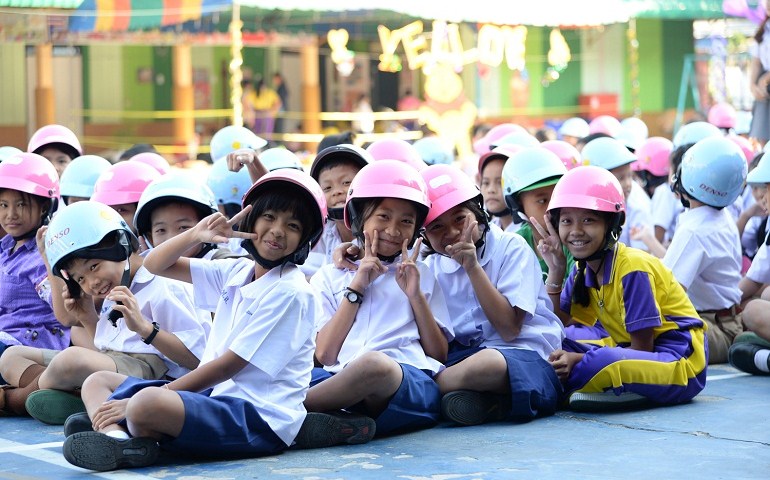 A poster displayed in Thai elementary schools to encourage students to wear bike helmets (left). Thai children don bike helmets donated by Denso (right) as part of the company’s local traffic safety program. (Courtesy of Denso)